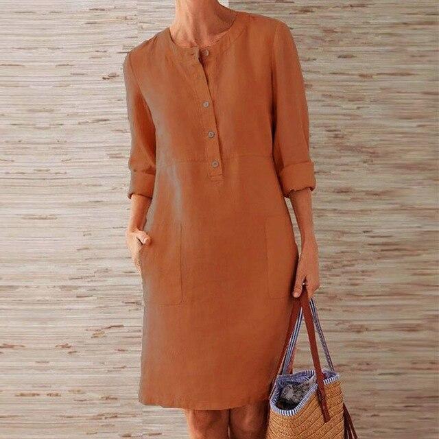 Donna con abito marrone a maniche lunghe, tasche frontali, tiene una borsa di paglia. Sfondo in legno chiaro. Moda casual elegante.
