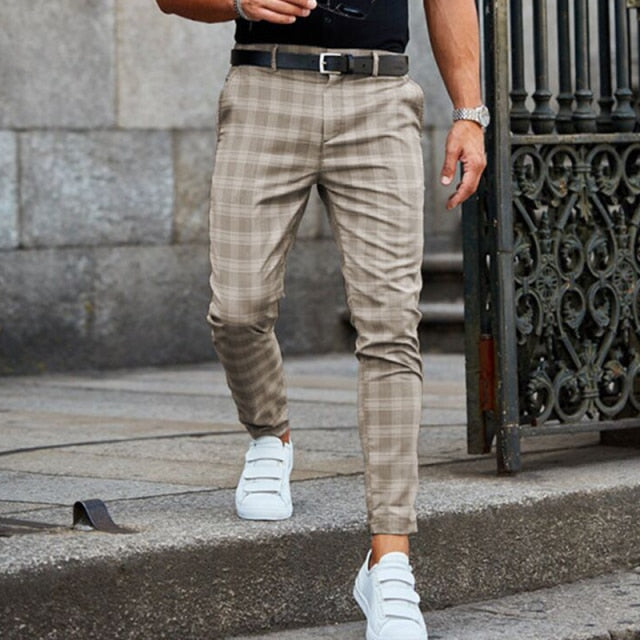 Uomo con pantaloni a quadri beige, cintura nera e scarpe da ginnastica bianche, cammina su marciapiede urbano. Moda maschile casual elegante.