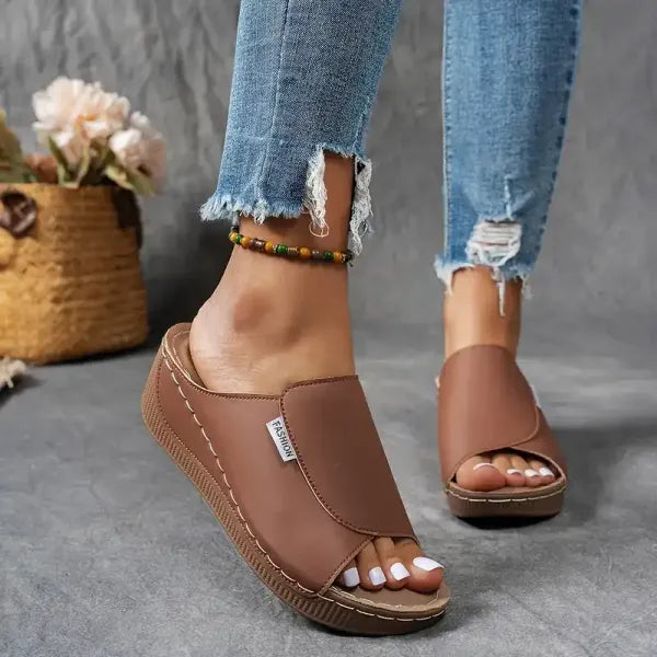 Sandali marroni in pelle con zeppa, indossati con jeans strappati. Sfondo con cesto di fiori. Scarpe estive eleganti e comode per donna.