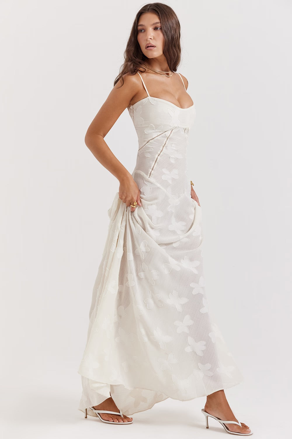 Donna in abito lungo bianco con motivo floreale, spalline sottili, sandali con tacco, posa elegante. Moda femminile, abbigliamento estivo chic.