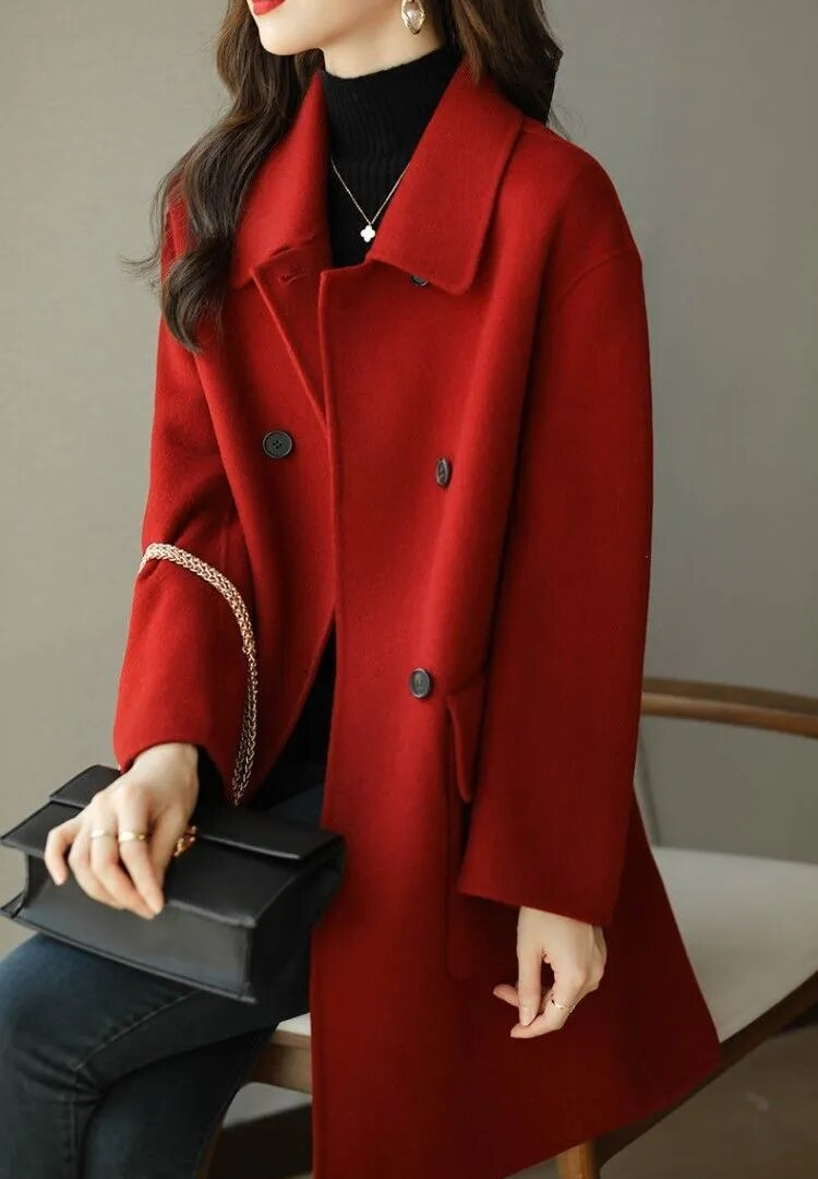 Donna con cappotto rosso elegante, seduta su sedia, indossa maglione nero e tiene borsa nera. Moda autunnale, stile chic, abbigliamento femminile.