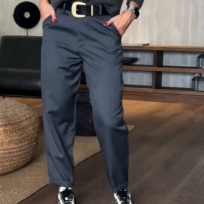 Pantaloni a righe grigie eleganti, cintura nera con fibbia dorata, scarpe bicolore, stile moderno, pavimento in legno, arredamento minimalista.