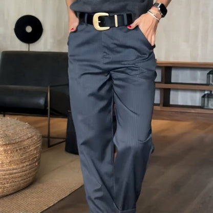 Donna in pantaloni eleganti grigi con cintura nera e fibbia dorata, mani in tasca. Arredamento moderno sullo sfondo. Moda femminile contemporanea.