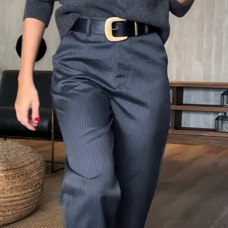 Donna in pantaloni a righe blu scuro, cintura nera con fibbia dorata, maglione grigio. Interno moderno con pavimento in legno. Moda autunnale elegante.