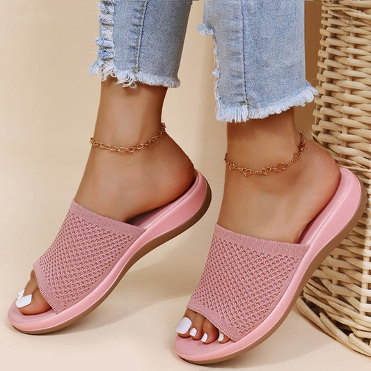 Sandali rosa da donna con suola piatta, indossati con jeans strappati. Design intrecciato, stile casual estivo. Accessoriati con cavigliere dorate.