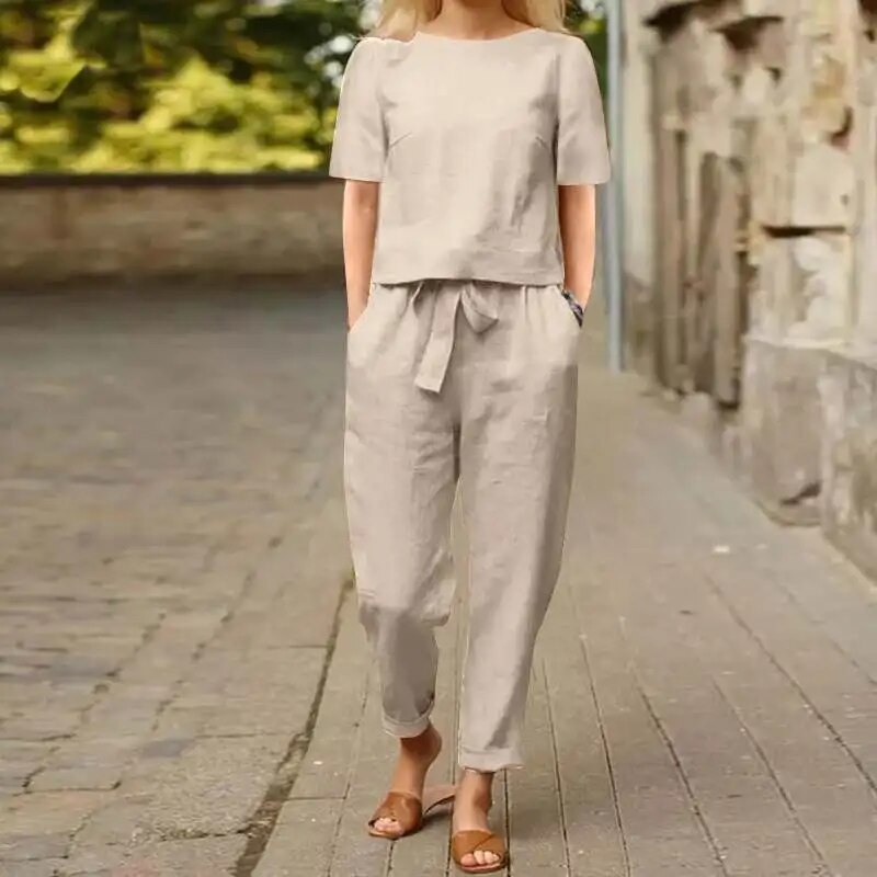 Donna con completo beige in lino, camicia e pantaloni, cammina su strada lastricata. Moda estiva casual, abbigliamento leggero e comodo.